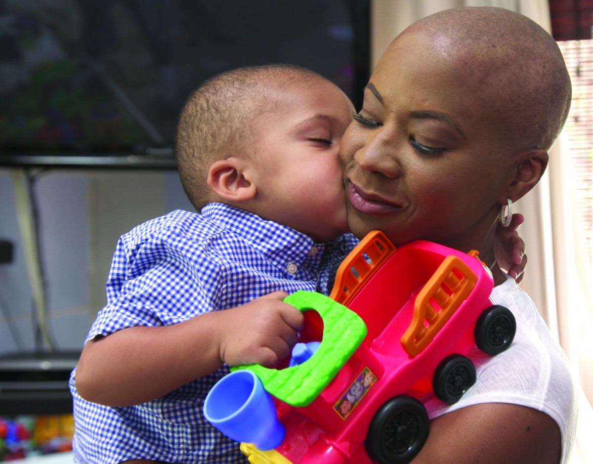 woman holding her child and his toy, while he kisses her on the cheak.