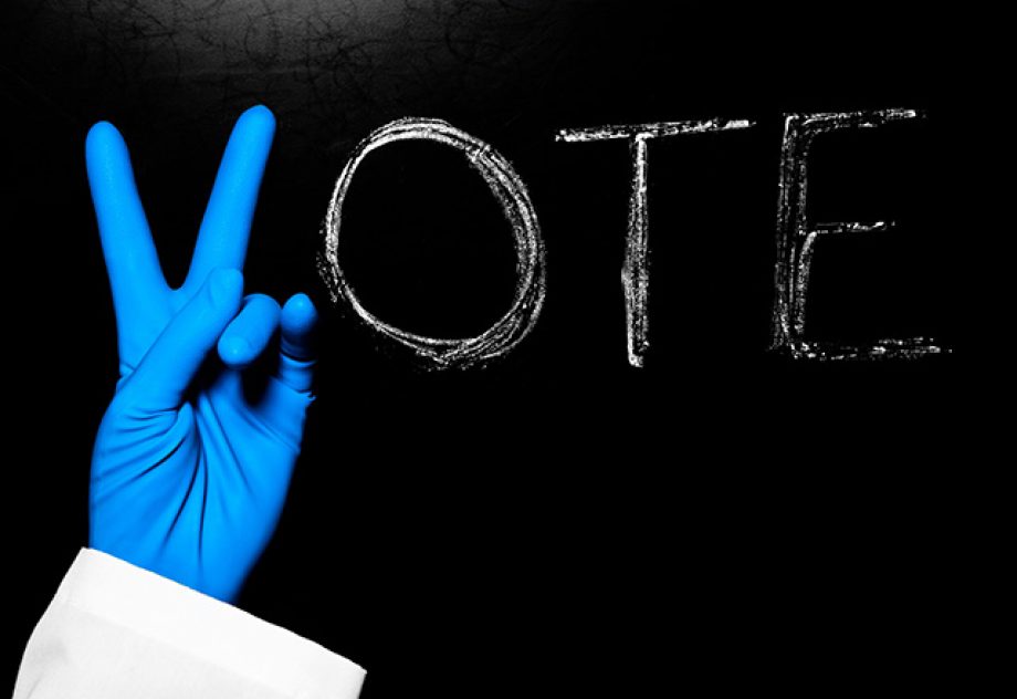 The word "Vote" is written on a blackboard, with a blue latex-gloved hand making the letter V.