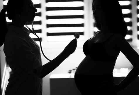 Silhouette of a female doctor holding a stethoscope to a pregnant woman's chest.