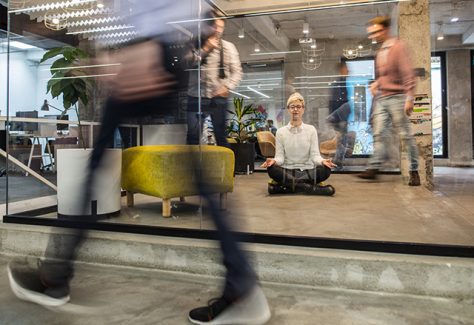 Young businesswoman meditating on the floor at her office while colleagues are walking in blurred motion. The view is through the glass.