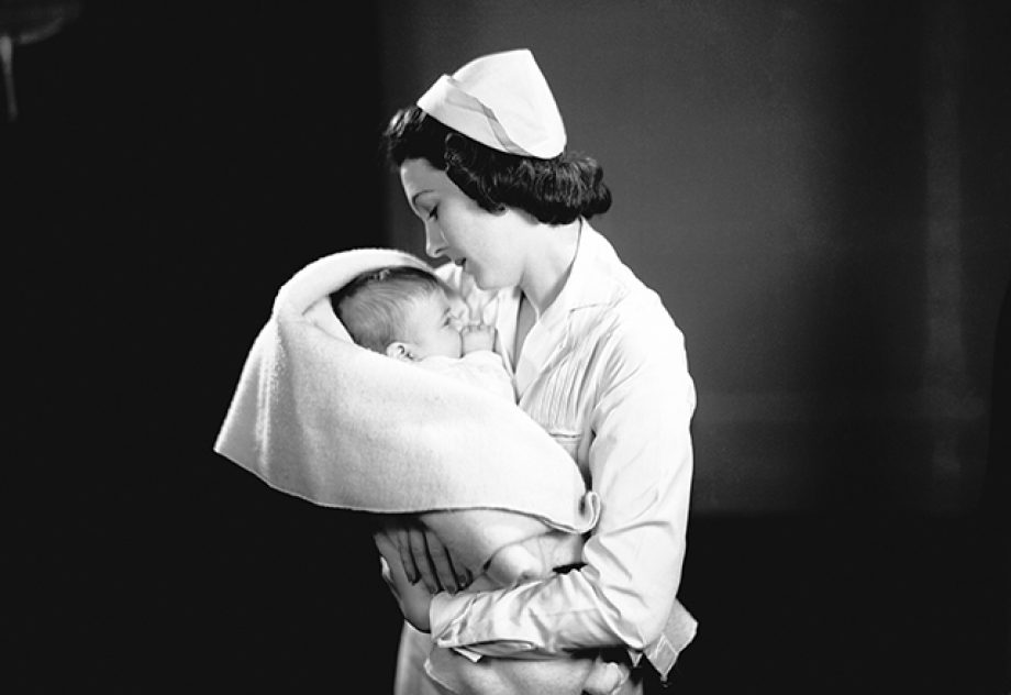 A 1950s-era nurse holds a baby, swaddled in a blanket.