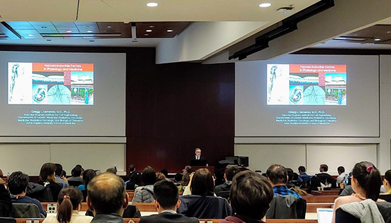 Dr. Semenza speaks in front of a full auditorium, with two screens displaying his presentation behind him.