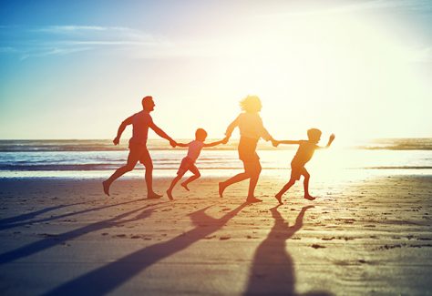A carefree family of four enjoy a run on the beach, with the sun behind them.