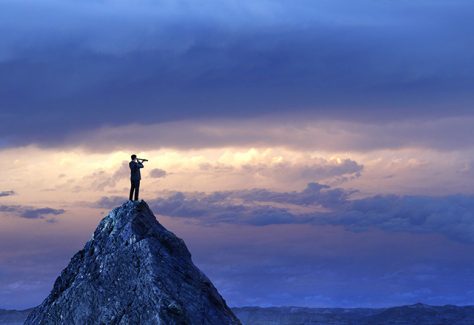 A silhouetted businessman stands on top of a mountain peak and looks into the distance with a spyglass in front of a dramatic sunset in the distance.