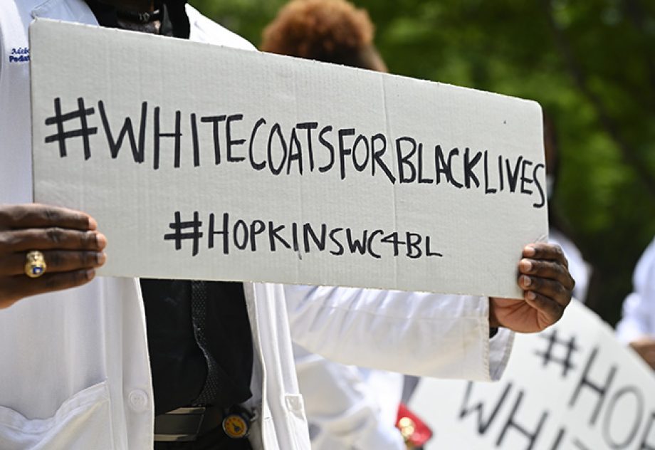 A Black member of Hopkins staff holds a sign that says, "White Coats for Black Lives."