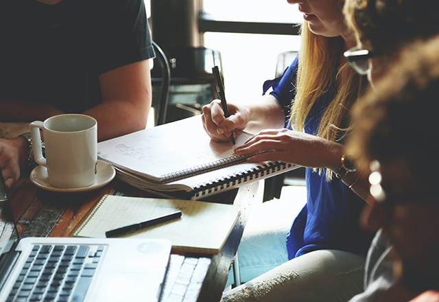 A group of people sit at a table in a coffeeshop, with a laptop and notebook.