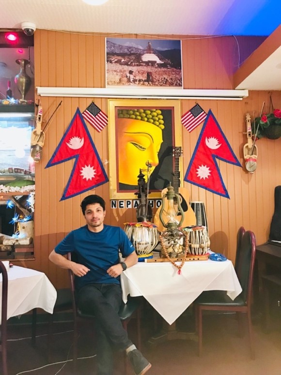 Surya sits at a table; there are traditional Nepalese instruments on the table. On the walls are posters and flags of Nepal.