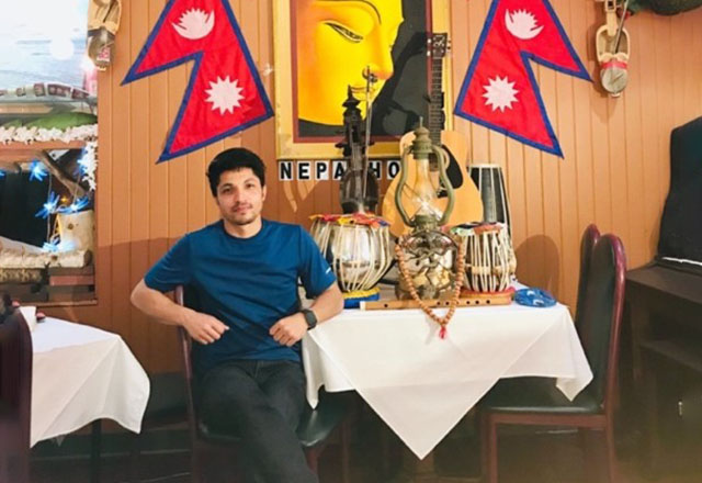 Surya sits at a table; there are traditional Nepalese instruments on the table. On the walls are posters and flags of Nepal.