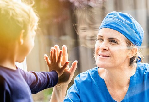 A healthcare worker presses her hand to a glass window, as a young boy holds his hand up on the other side.