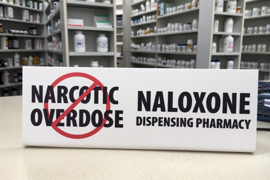 Banner on pharmacy counter against narcotic overdose, with "naloxone dispensing pharmacy" listed.