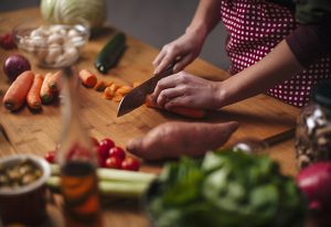 Hands of young woman as she chopping carrots with big knife with various vegetables around on kitchen counter