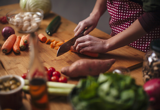 Hands of young woman as she chopping carrots with big knife with various vegetables around on kitchen counter