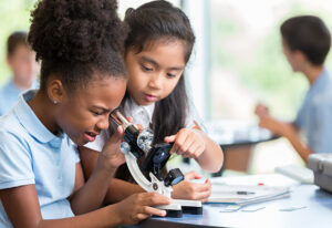 Focused African American and Asian schoolgirls help one another with their assignment in science class at a STEM elementary school. The African American girl is looking at something with a microscope.