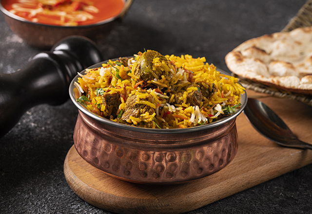 Chicken and meat dhum biriyani using jeera rice and spices arranged in earthen ware with raitha and lemon pickle on background.