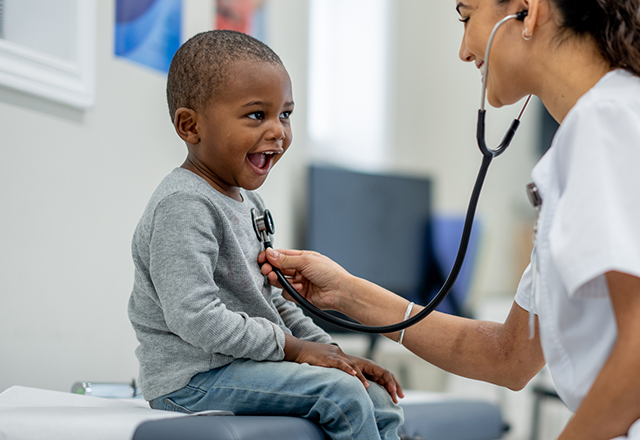A young boy sits up on an exam table as a female Pediatrician preforms a check-up on him. The boy is dressed casually and smiling as the doctor listens to his heart.